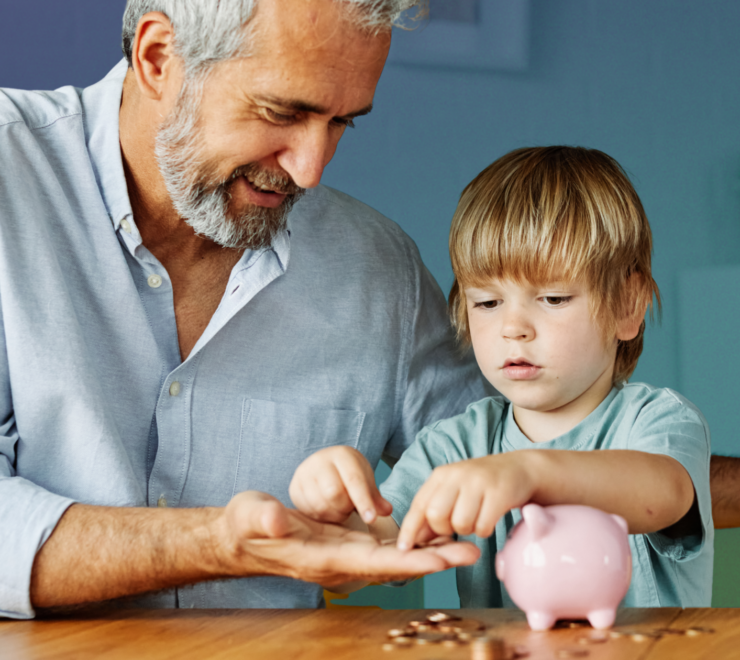 A smiling grandfather and young grandson sitting at a table together, counting coins from a pink piggy bank. This image represents the intergenerational wealth-building potential of Trump Accounts and long-term financial planning for children.