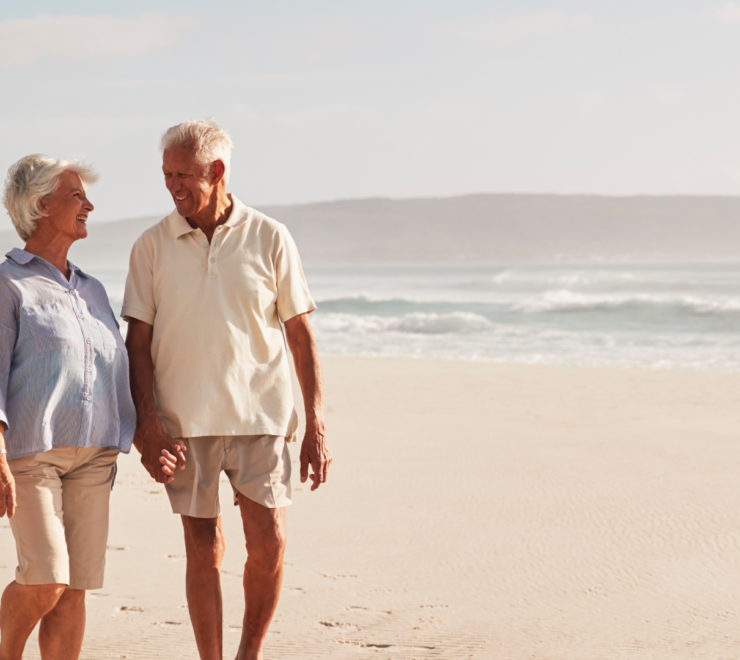 Two retirees walking hand in hand on a beach, smiling at each other on a sunny day.